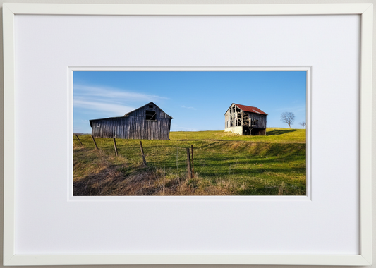 Framed photograph of two old barns on a grassy hill with a clear blue sky in iowa