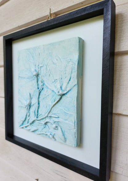 Righthand view of a white cast magnolia flowers color washed in blue in a black frame with a white backerboard.