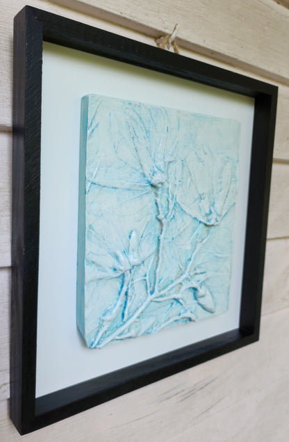 Lefthand view of a white cast magnolia flowers color washed in blue in a black frame with a white backerboard.