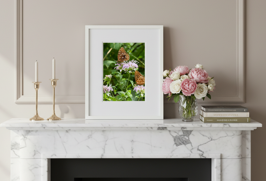 Framed photograph of two butterflies on flowers on a marble fireplace mantel with candles and books.