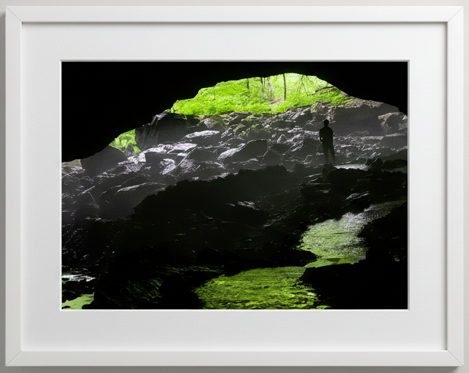 Phot print of a Person standing inside a cave with green light filtering through the forest outside at maquoketa caves state park in iowa