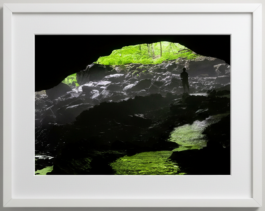 Phot print of a Person standing inside a cave with green light filtering through the forest outside at maquoketa caves state park in iowa